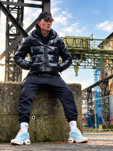 Sven in Nike TN1 "Marseille", schwarzer Jogger und Glanz-Pufferjacket im Industriegebiet